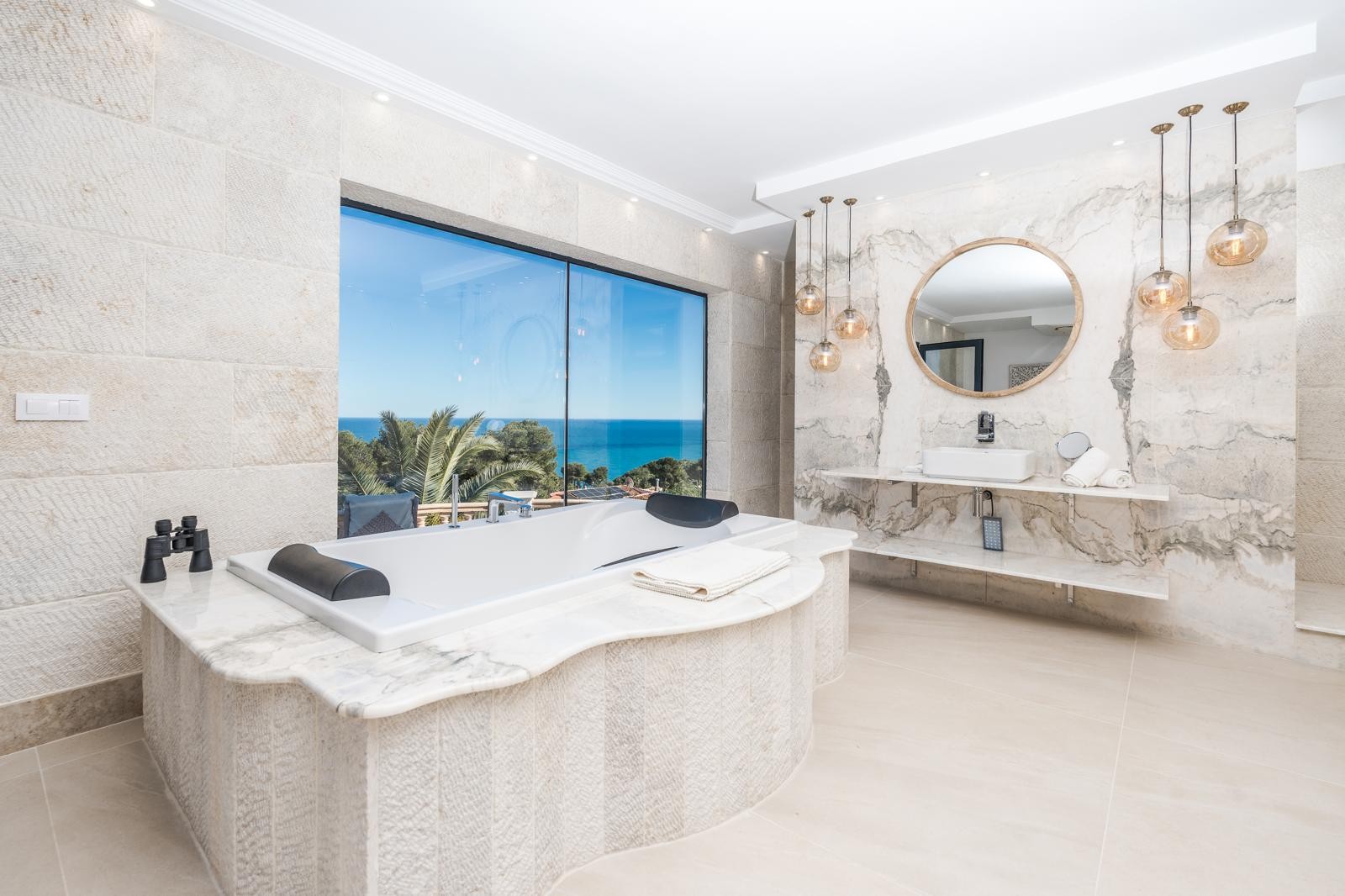 Baño de la suite principal con bañera encastrada en mármol frente a un gran ventanal con vistas al mar, paredes de piedra natural y lavabo sobre encimera de mármol.