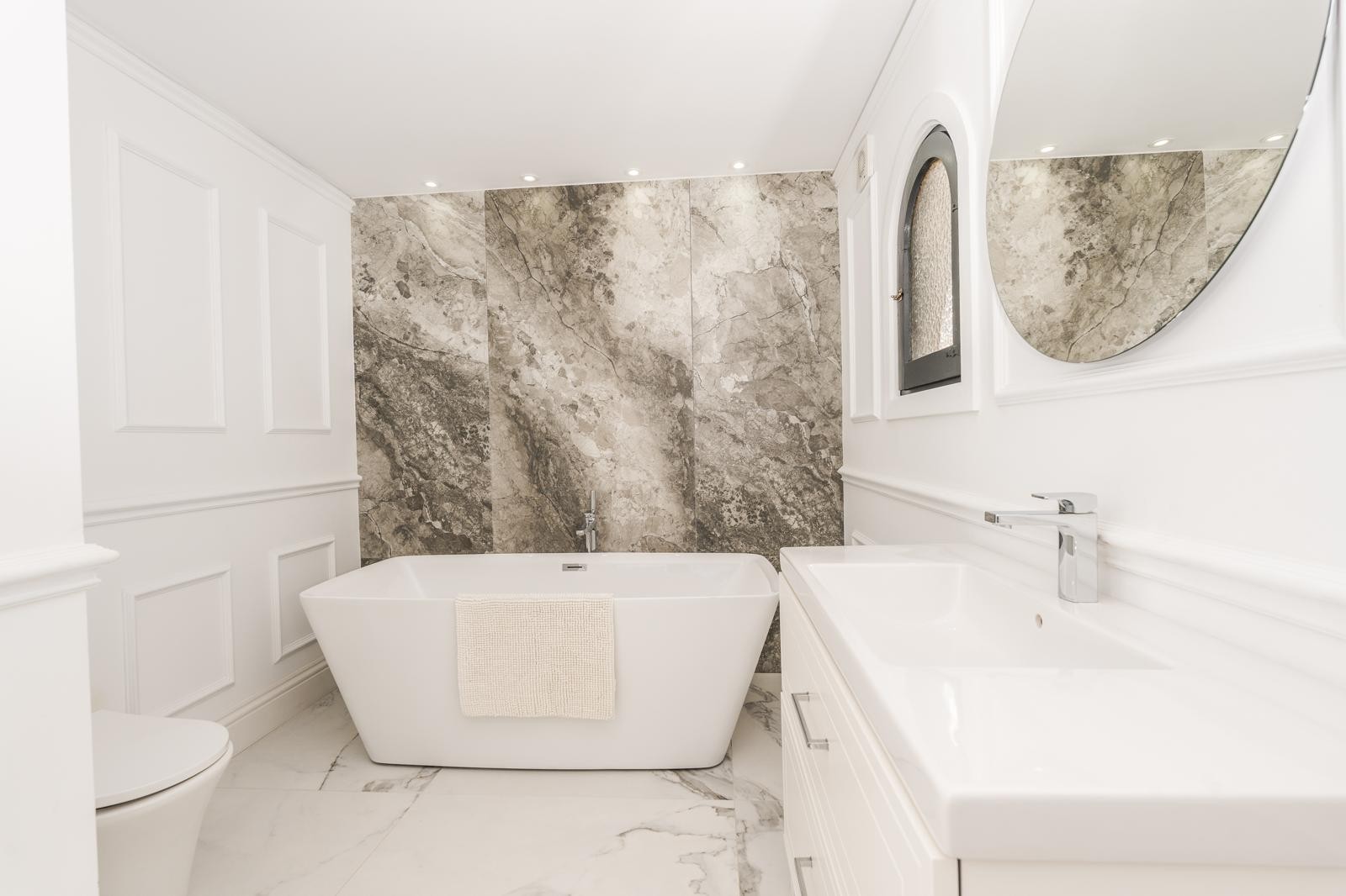Baño de la suite principal con bañera exenta, pared de mármol veteado y molduras decorativas. Incluye lavabo integrado, espejo circular y focos empotrados.