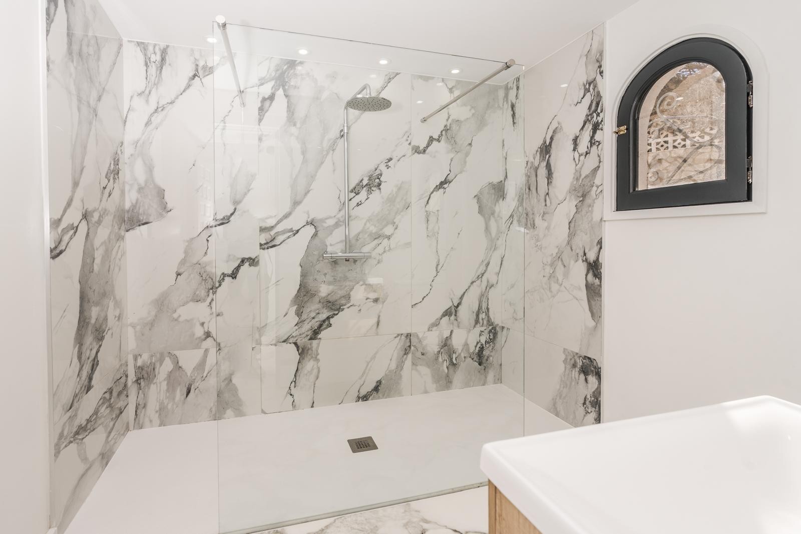 Baño moderno con ducha walk-in revestida en mármol de gran formato, mampara de cristal fija y ventana de arco con marco negro que aporta luz natural.