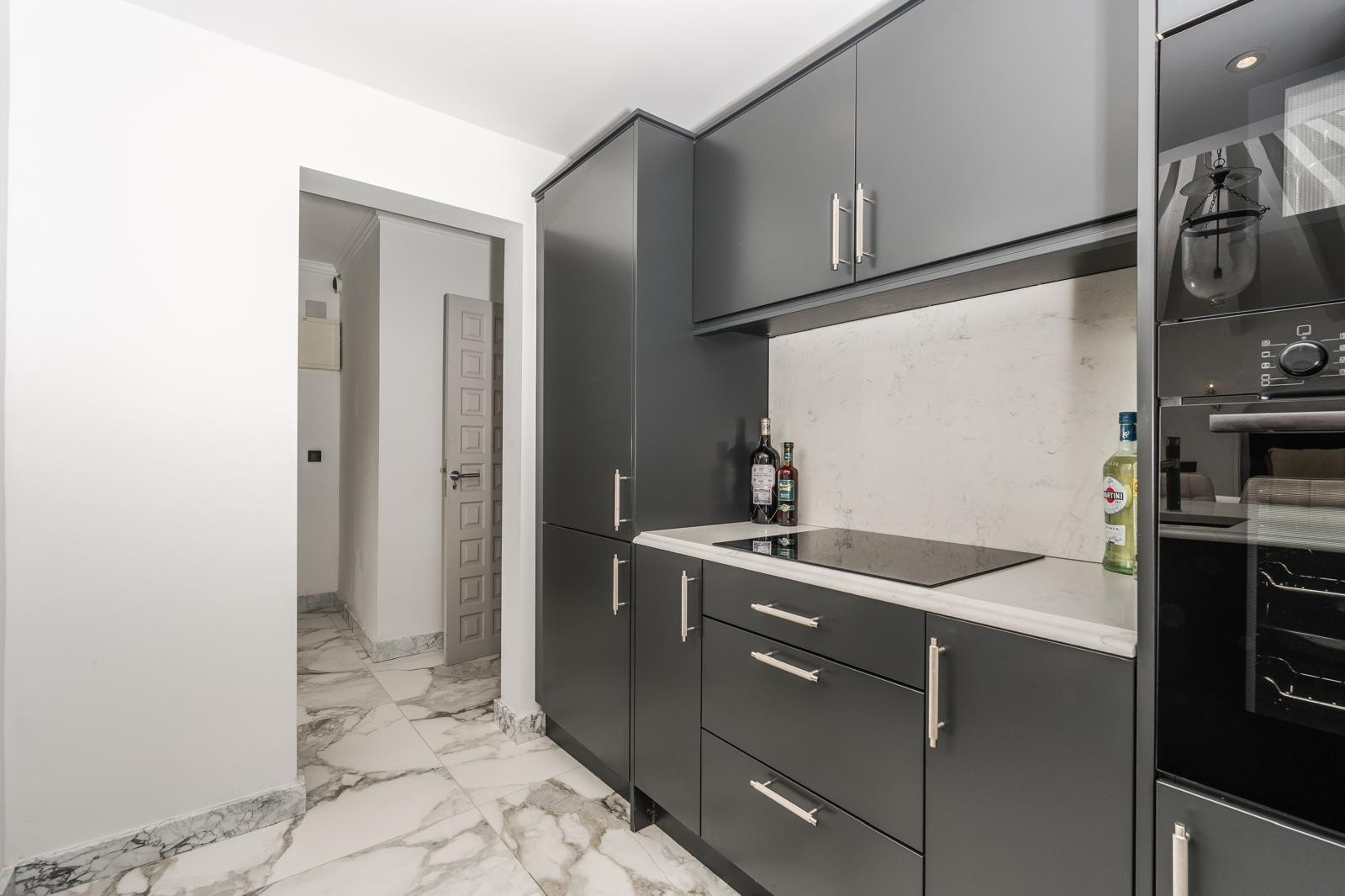 Cocina moderna con armarios en gris grafito, encimera de mármol blanco y placa de inducción integrada. El suelo de mármol veteado conecta con el pasillo y la zona de electrodomésticos.