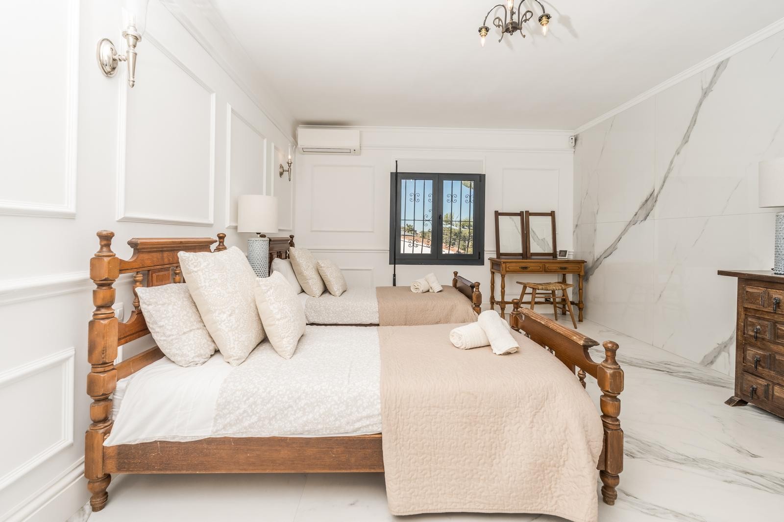 Dormitorio con dos camas de madera, paredes con molduras clásicas y un revestimiento de mármol veteado. Incluye tocador rústico, suelos de mármol y aire acondicionado.