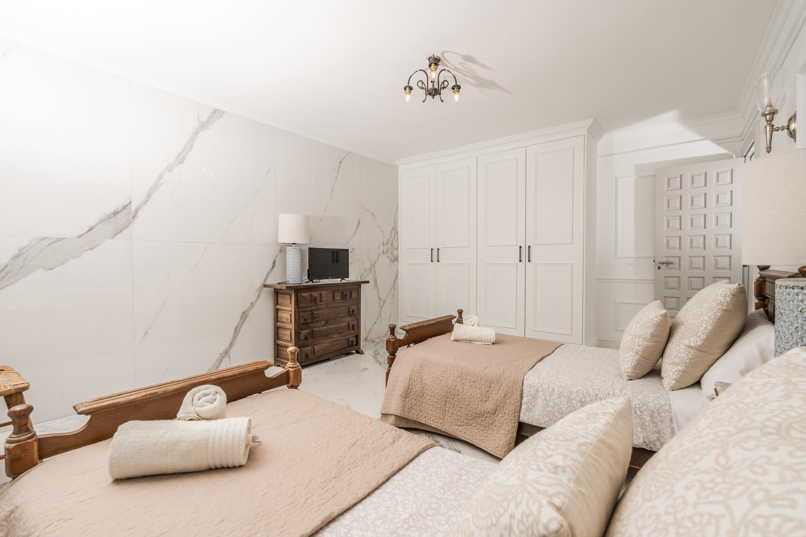 Dormitorio con dos camas individuales de madera, pared revestida en mármol veteado y armarios empotrados blancos. Incluye cómoda rústica y suelos de piedra clara.
