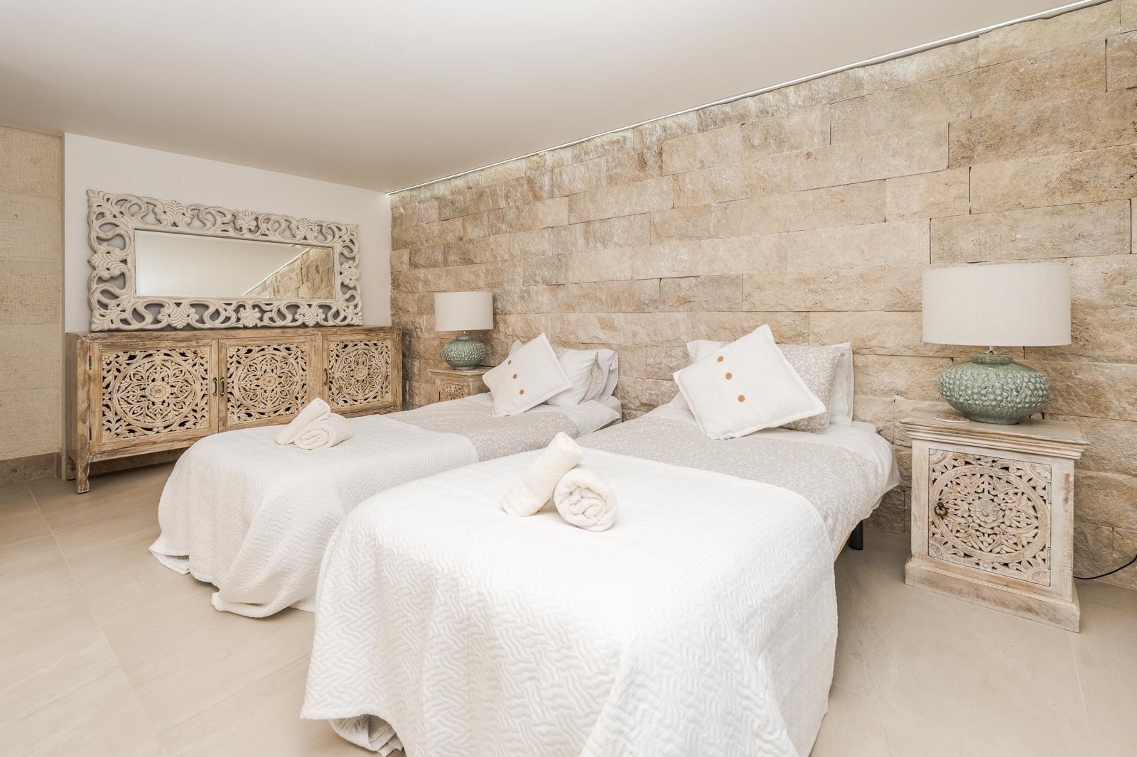 Dormitorio con pared de piedra natural, dos camas individuales y mobiliario de madera tallada de estilo mediterráneo. Incluye iluminación ambiental y suelos de tonos claros.