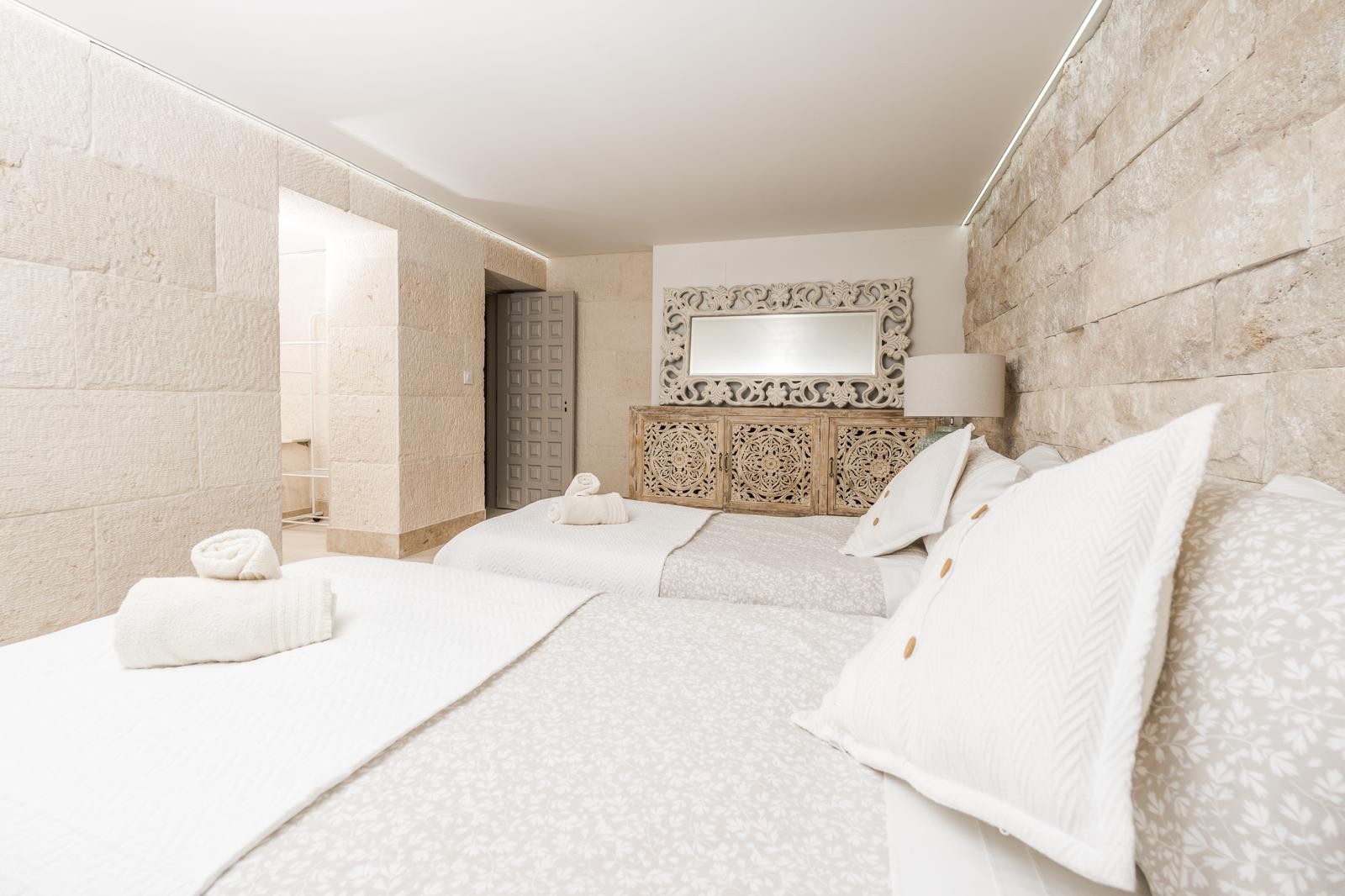 Dormitorio con paredes de piedra natural, dos camas individuales y mobiliario de madera tallada. Incluye iluminación LED perimetral y acceso a zona de vestidor.