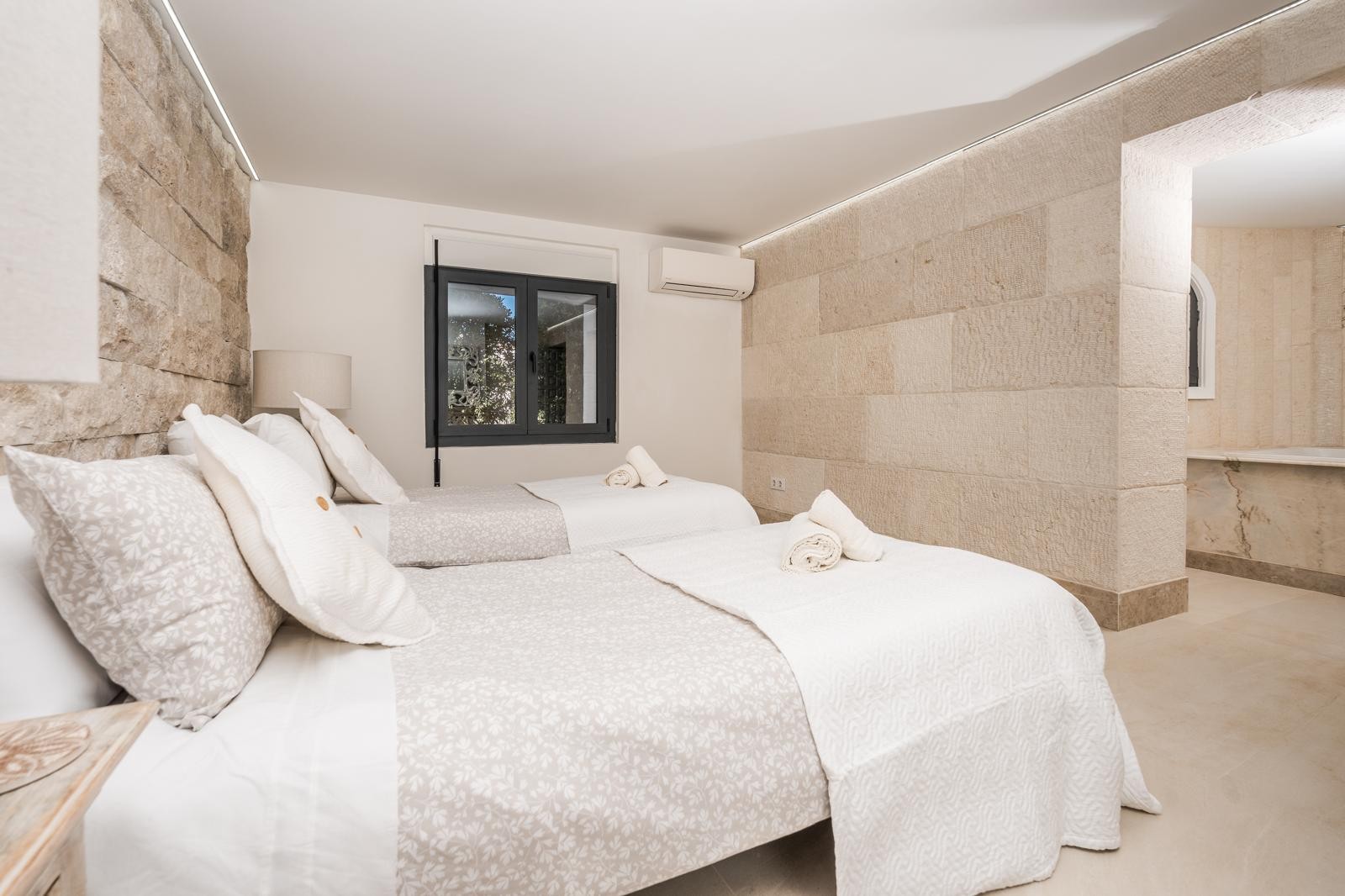 Dormitorio con paredes de piedra natural, iluminación LED indirecta y dos camas individuales. Incluye aire acondicionado y acceso a un baño con acabados en mármol.