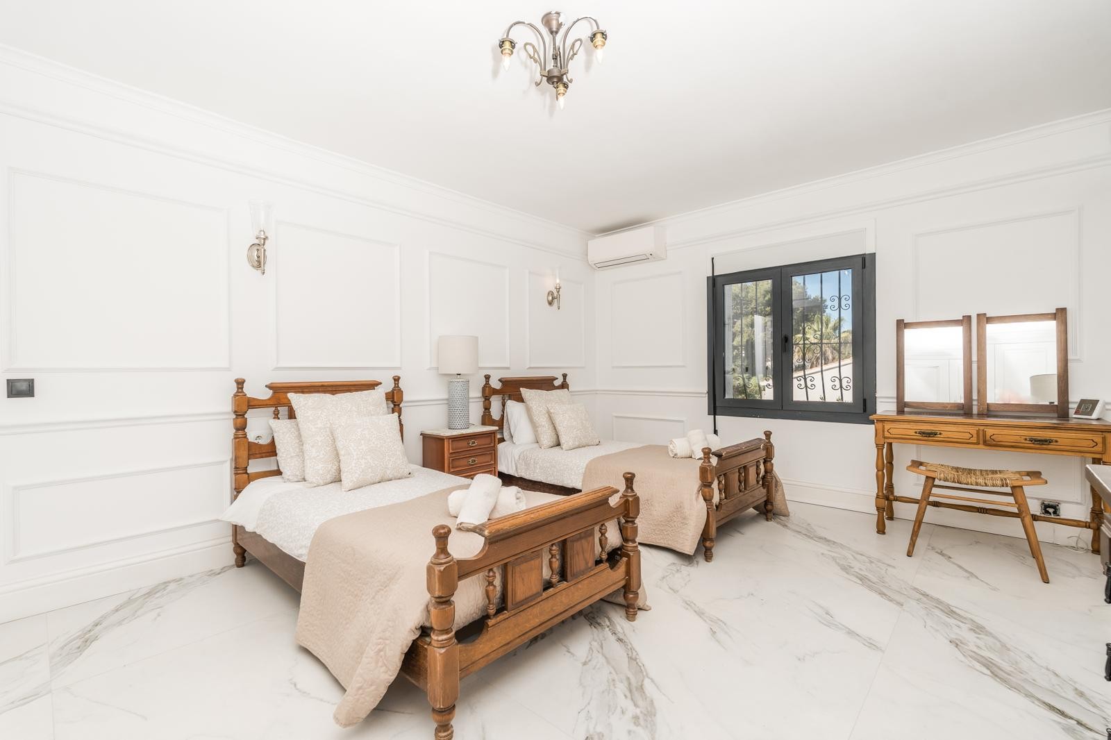 Dormitorio luminoso con dos camas individuales de madera tallada, suelos de mármol veteado y paredes con molduras clásicas. Incluye tocador de madera y aire acondicionado split.