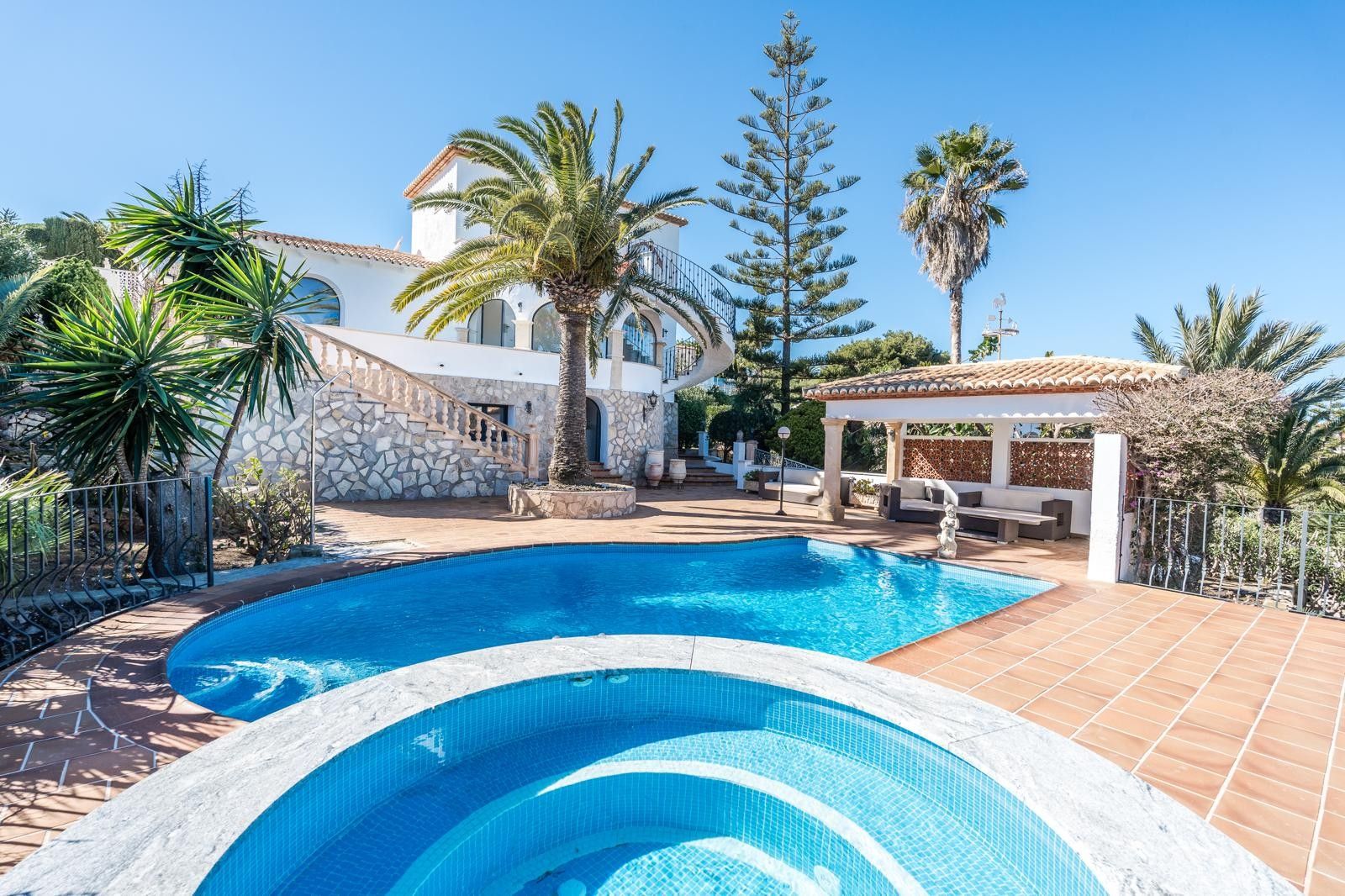 Exterior de la villa con piscina renovada, jacuzzi circular en primer plano y porche cubierto. Destaca la fachada blanca con zócalo de piedra y palmeras mediterráneas maduras.