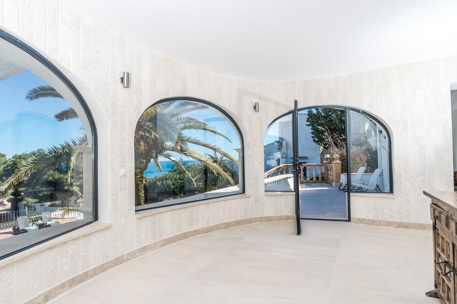 Salón de diseño circular con paredes revestidas en piedra natural y grandes ventanales en arco que ofrecen vistas al mar y acceso directo a la terraza exterior.