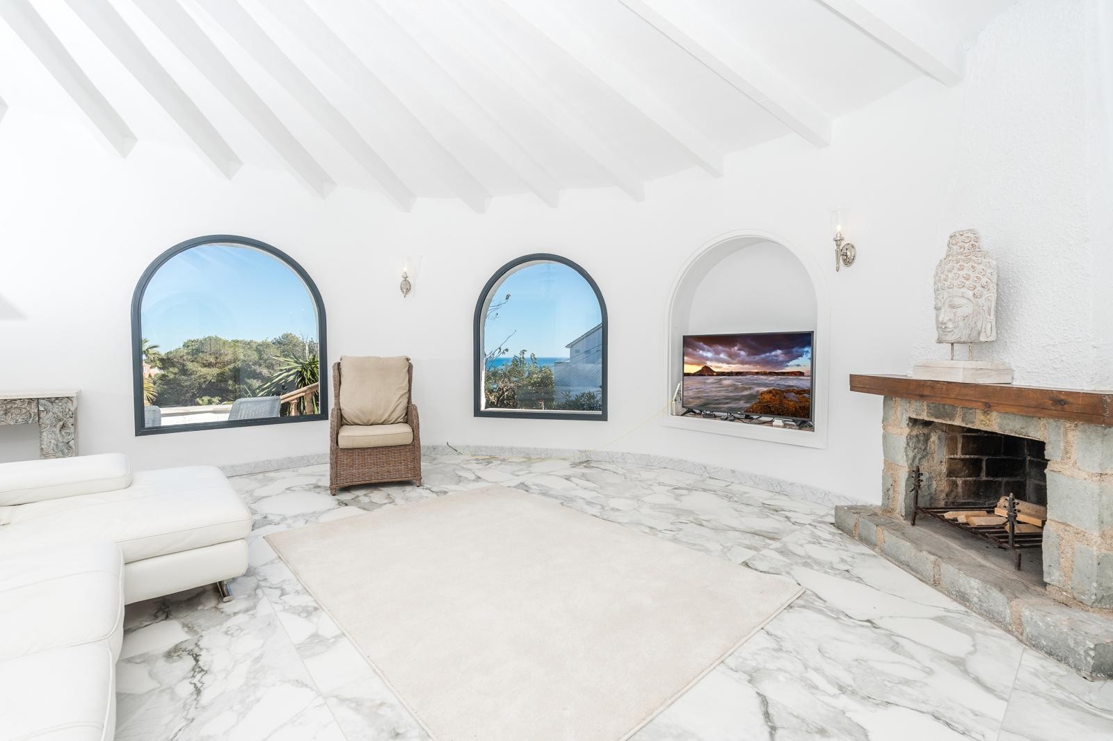 Salón de planta circular con techos de vigas blancas, suelo de mármol y chimenea de piedra. Ventanales de arco ofrecen vistas al exterior y una luminosidad natural constante.