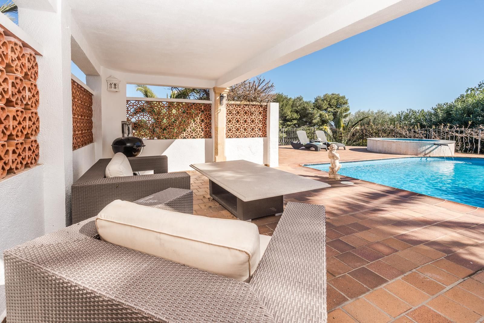 Terraza cubierta con mobiliario de ratán gris y mesa de piedra, situada junto a la piscina renovada y el jacuzzi exterior rodeado de vegetación mediterránea.