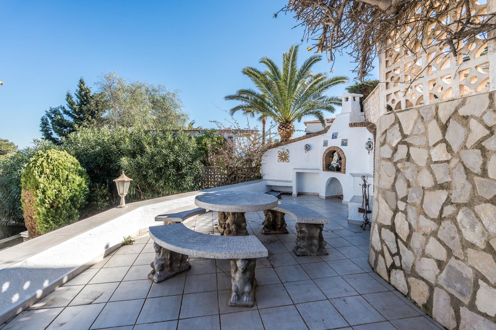 Terraza exterior con conjunto de mesa y bancos de piedra tallada, barbacoa de obra integrada y muros revestidos en piedra natural bajo un entorno mediterráneo.
