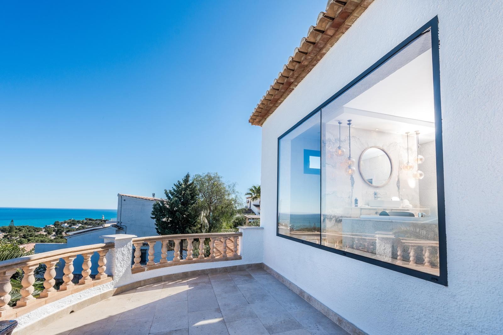 Terraza superior con balaustrada de piedra y vistas al mar, junto a un gran ventanal que muestra el baño de diseño con mármol y luminarias colgantes.