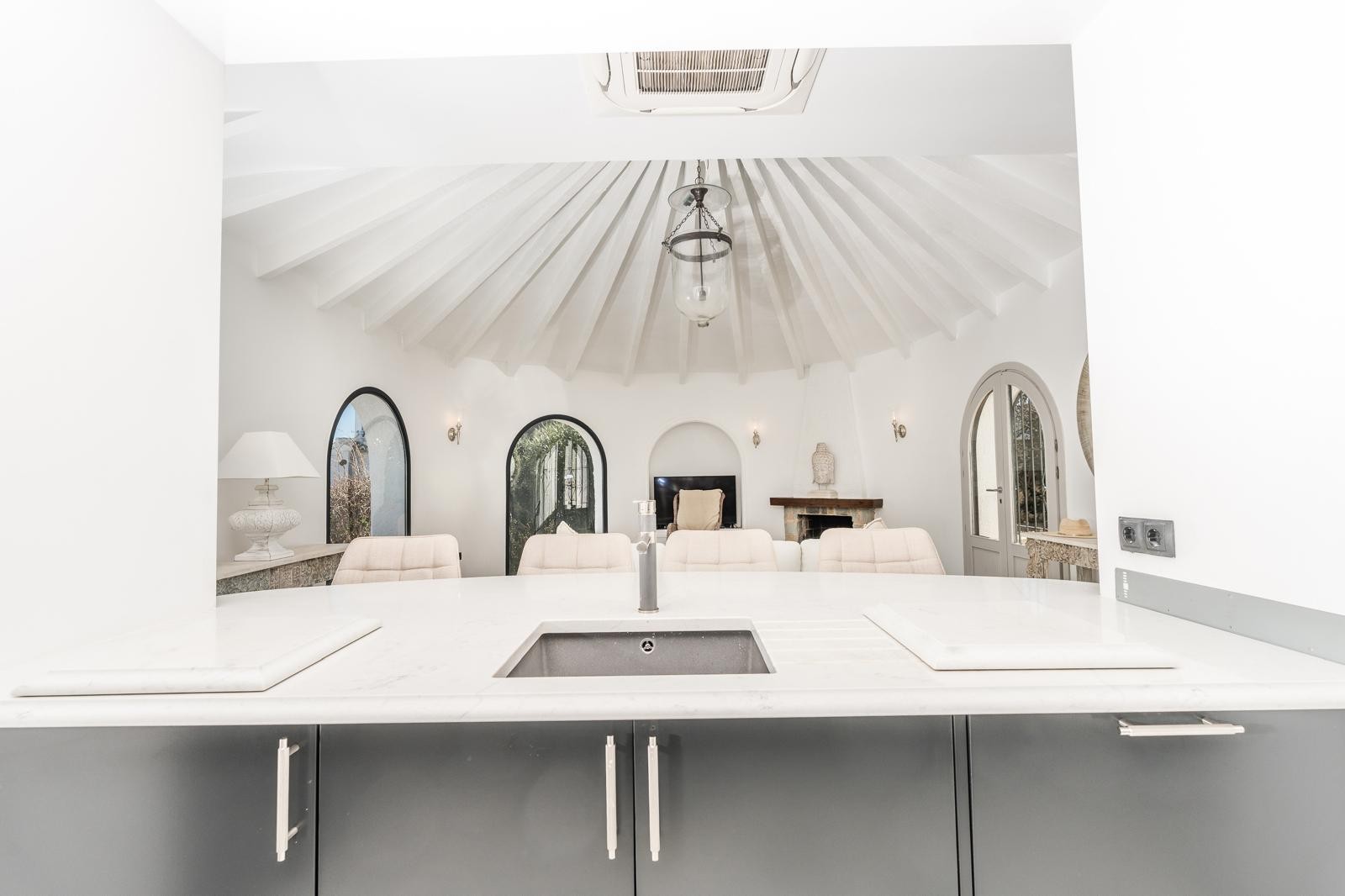Vista desde la cocina con encimera de mármol hacia el salón circular, destacando el techo abovedado con vigas blancas y la chimenea de piedra.