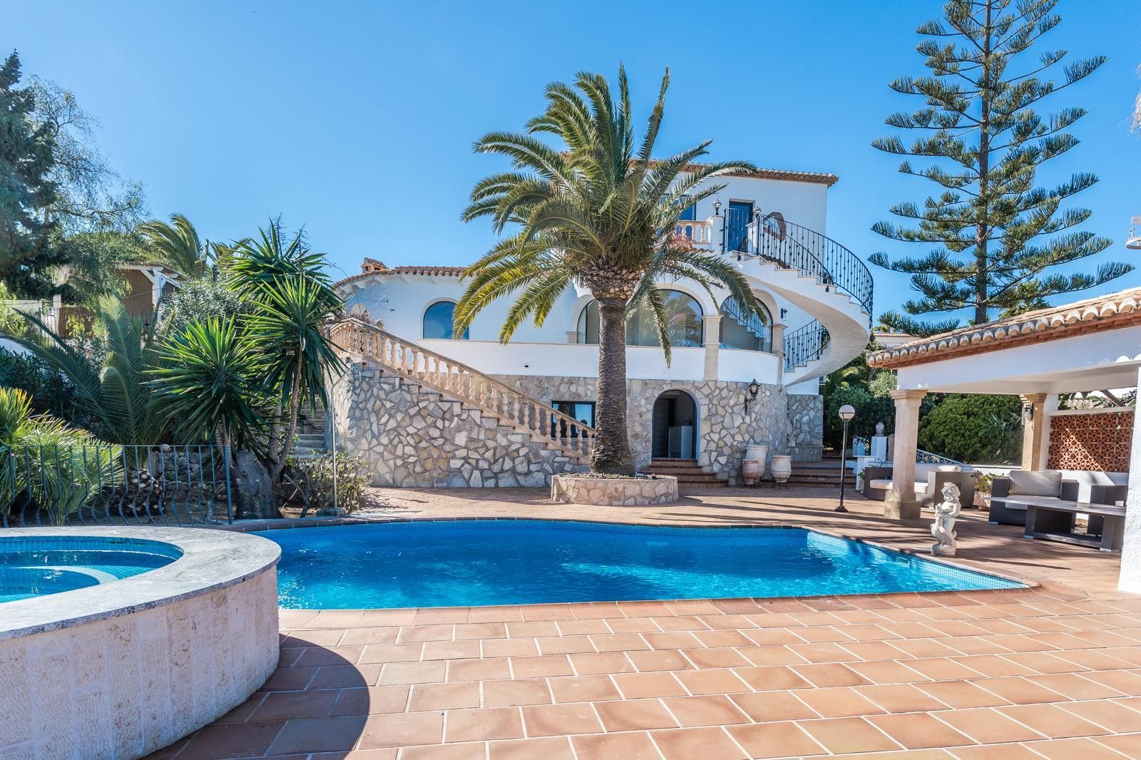 Vista exterior de la villa con piscina, jacuzzi circular y fachada de piedra natural. Incluye una escalera de caracol y jardín mediterráneo con palmeras bajo el sol.