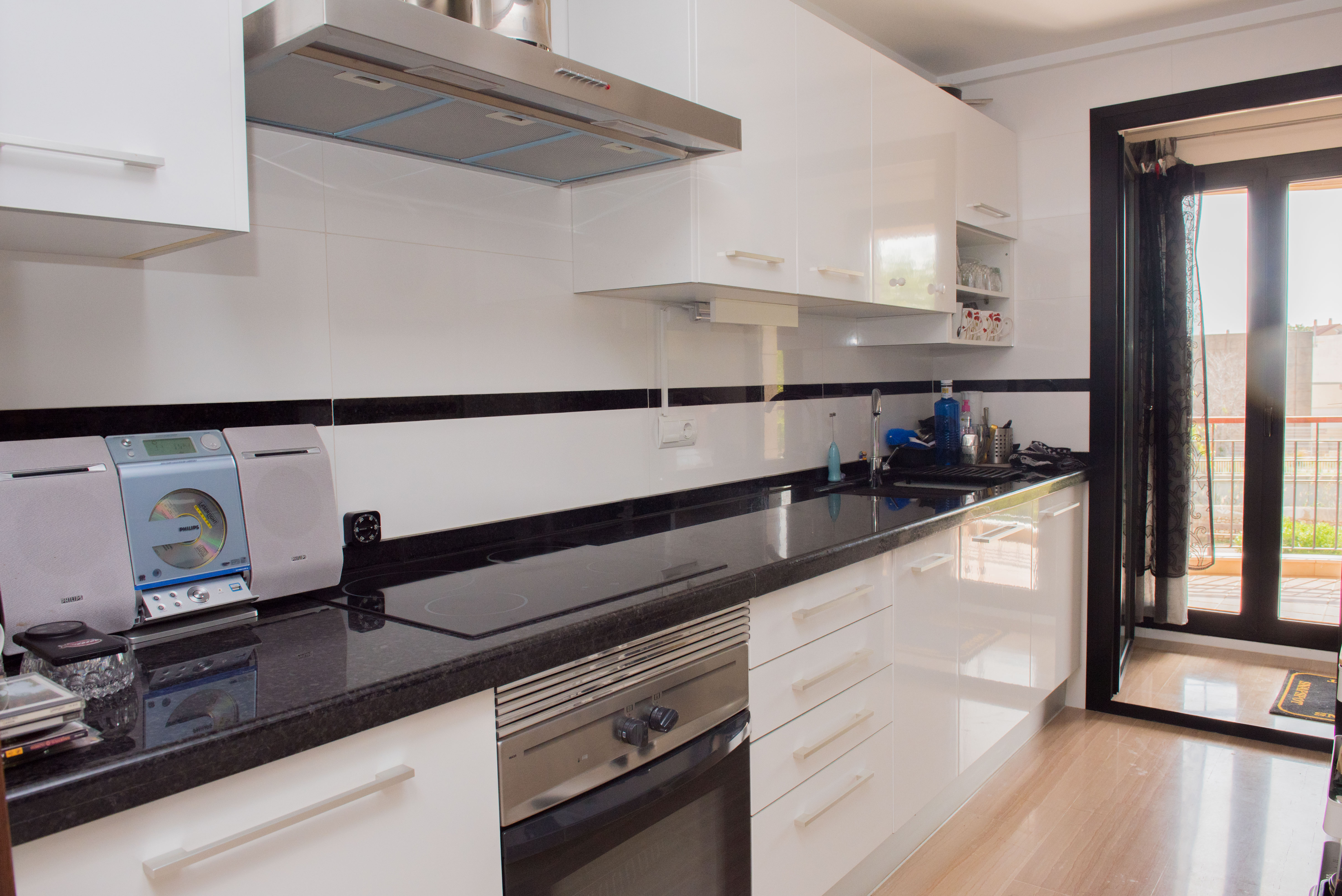 Cocina equipada con armarios blancos de alto brillo, encimera de granito negro y electrodomésticos integrados. Incluye acceso directo a un balcón exterior.