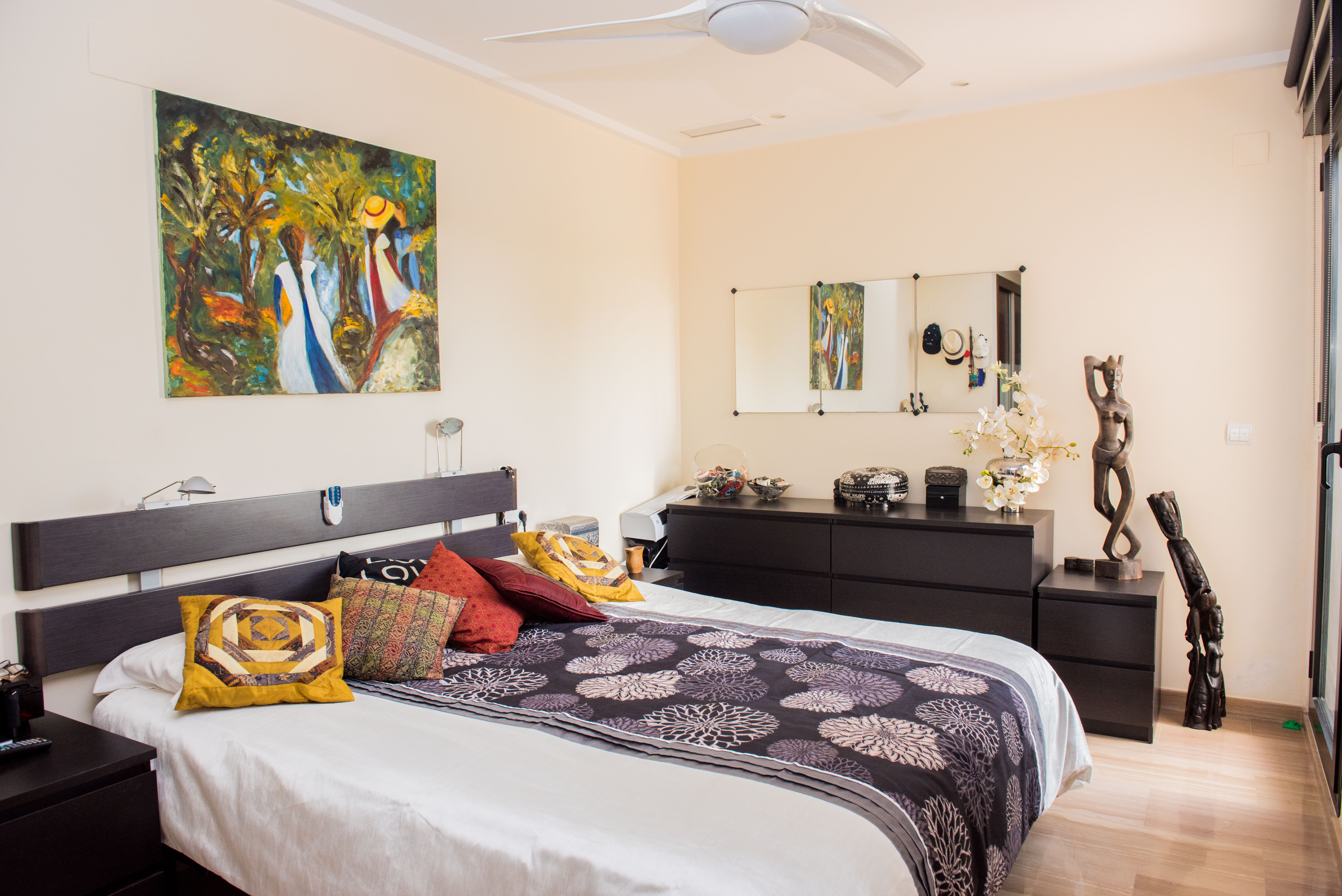 Dormitorio con cama doble, cabecero de madera oscura y ventilador de techo. Dispone de cómoda a juego, arte mural colorido y suelos de madera clara bajo luz natural.