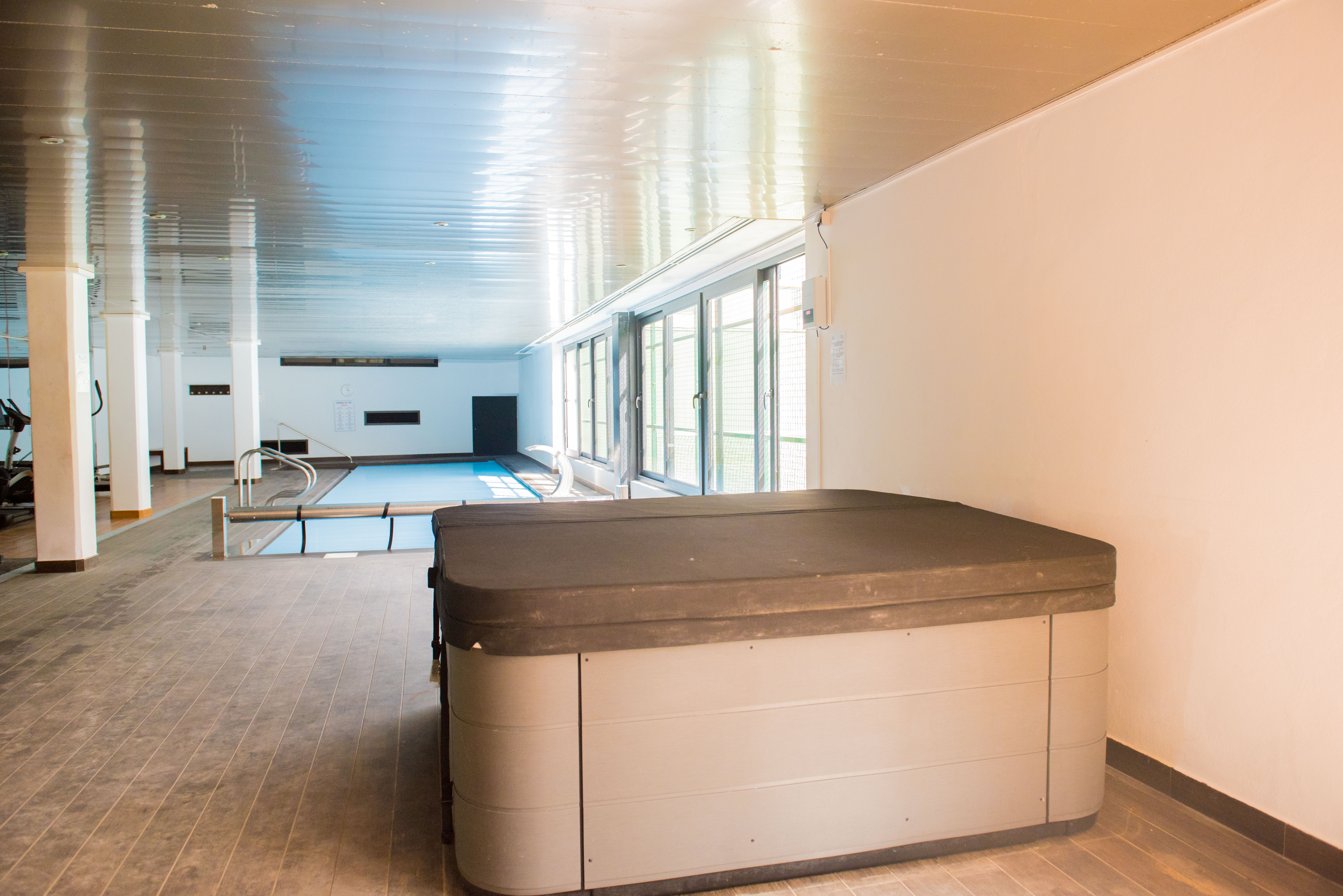 Zona de bienestar interior con jacuzzi cubierto, piscina y área de gimnasio, iluminada por amplios ventanales laterales y techo reflectante.