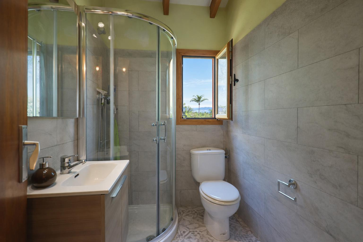 Baño funcional con ducha de cabina curva, paredes alicatadas en gris y vigas de madera. La ventana ofrece luz natural y vistas despejadas al entorno de Jávea.