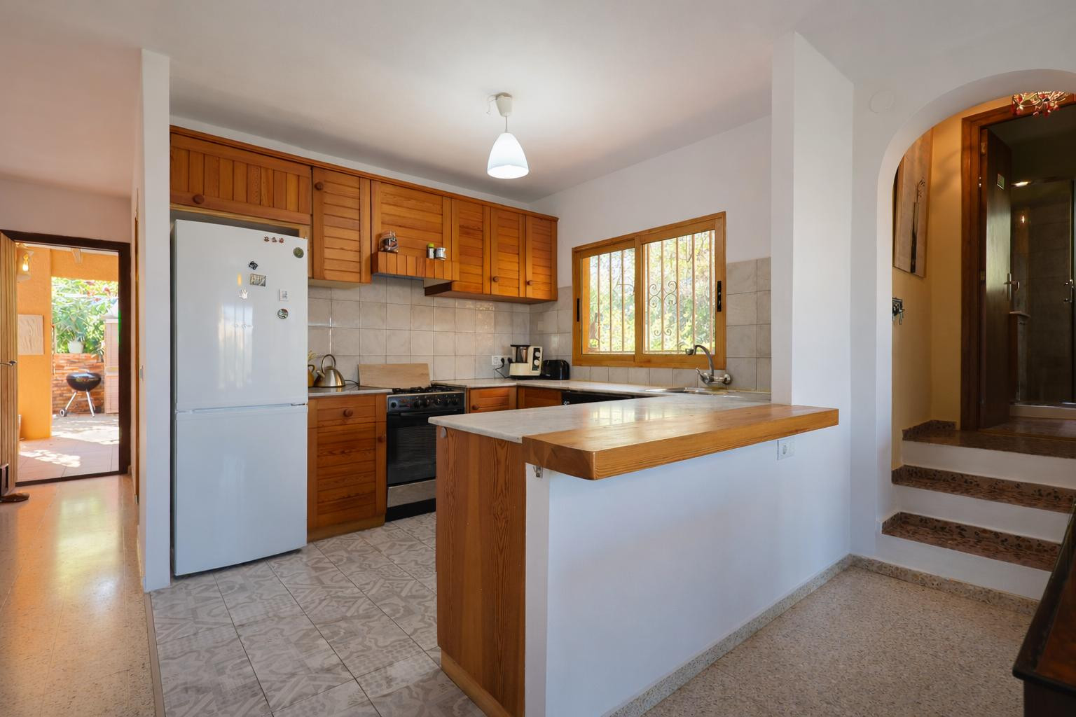 Cocina abierta con mobiliario de madera, encimera de mármol y barra integrada. Dispone de ventana al exterior, suelos cerámicos y conexión visual con la zona de barbacoa.