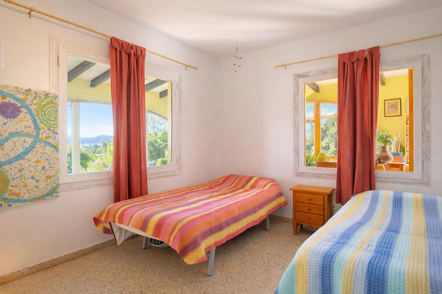 Dormitorio luminoso con dos camas individuales, mesita de noche de madera y ventanas de marco blanco que ofrecen vistas al paisaje y al interior de la vivienda.