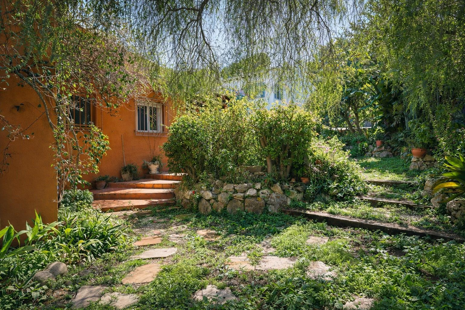Jardín mediterráneo con senderos de piedra y traviesas de madera, rodeado de vegetación madura y muros de piedra natural junto a la fachada terracota.