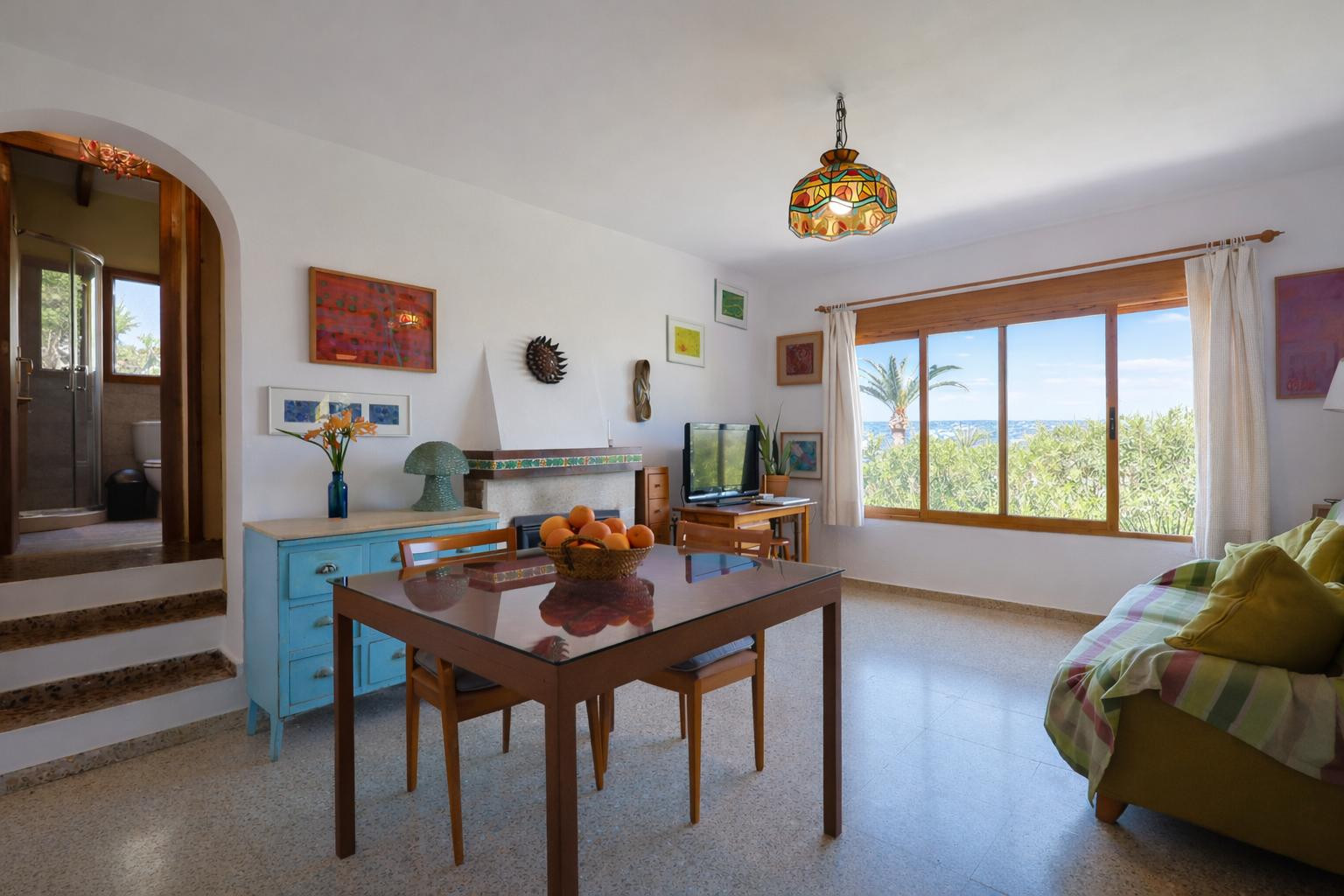 Salón comedor con chimenea, suelos de terrazo y ventanal con vistas al mar. El espacio conecta mediante un arco con un baño completo situado en un nivel superior.