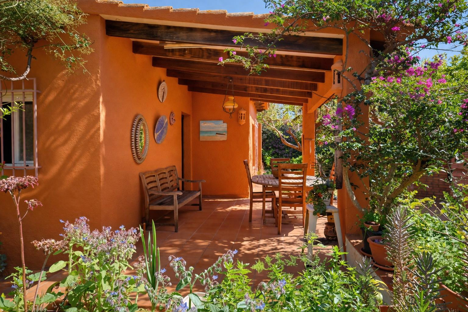 Terraza cubierta con vigas de madera vista, mobiliario de teca y suelos de barro, rodeada de vegetación mediterránea y buganvillas en flor.