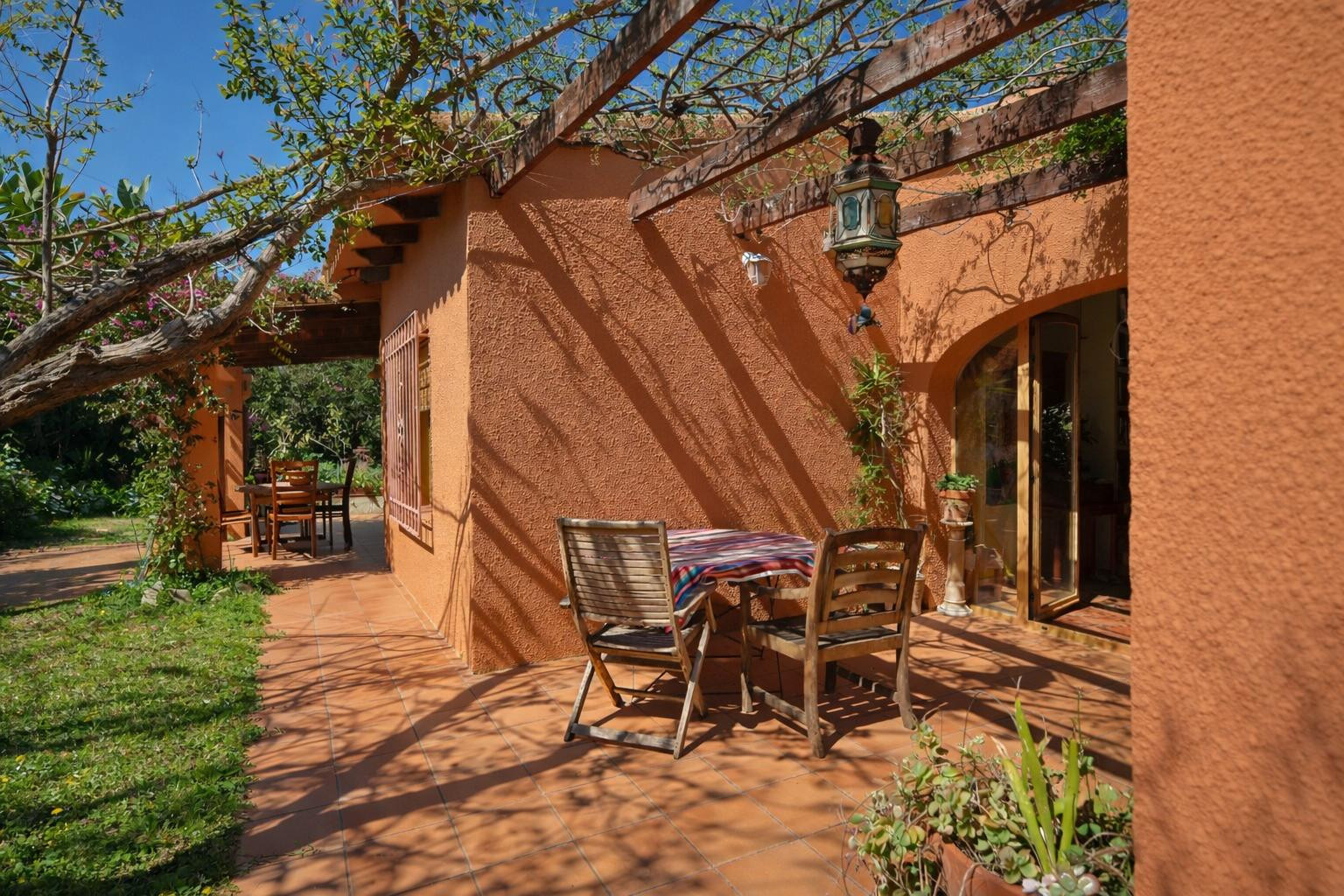 Terraza exterior con suelos de barro, pérgola de madera con vegetación y mobiliario de comedor al aire libre bajo una fachada de tono cálido.