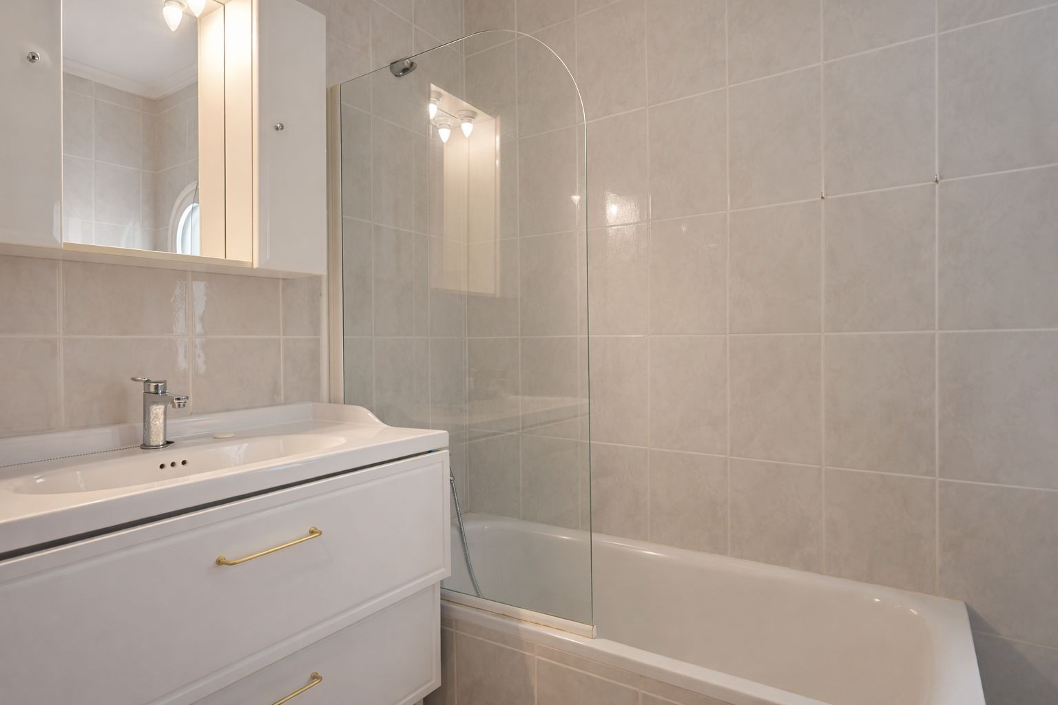 Baño completo con bañera y mampara de cristal curva, mueble de lavabo blanco con tiradores dorados y armario de espejo con iluminación superior.