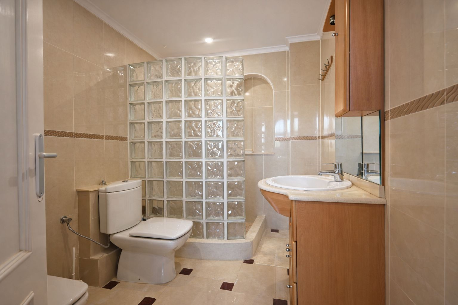 Baño completo con paredes alicatadas en tonos crema, ducha con mampara de bloques de vidrio y mueble de lavabo en madera con espejo integrado.