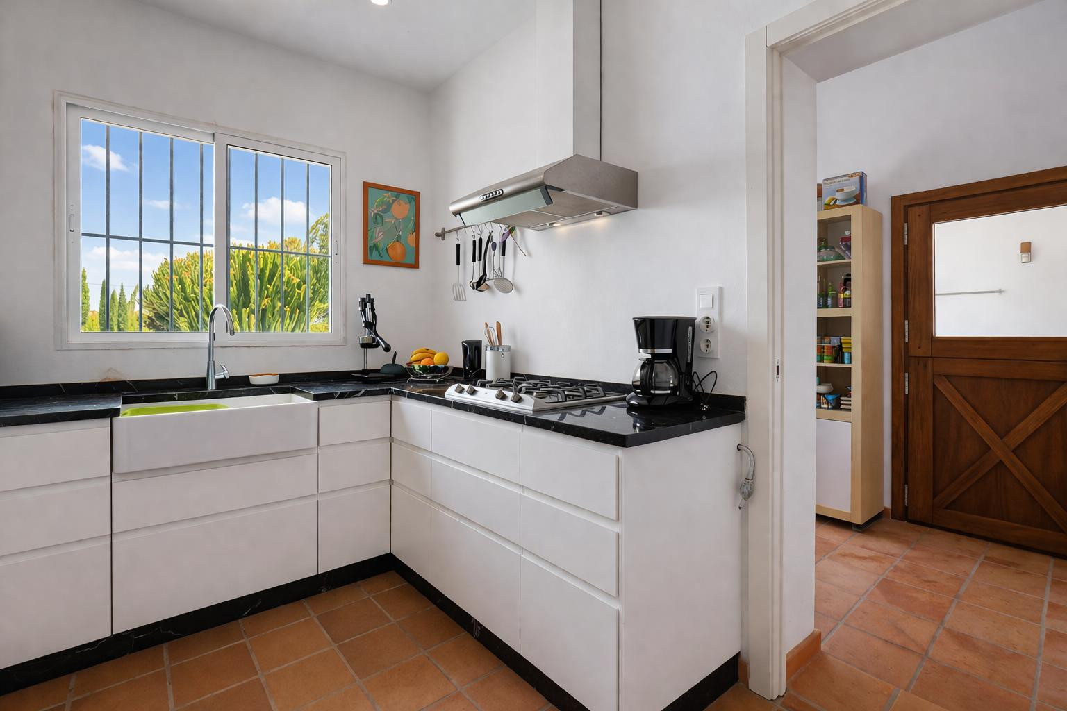 Cocina funcional con encimera de piedra negra, muebles blancos sin tiradores y fregadero cerámico. El suelo de terracota y la luz natural realzan el espacio de trabajo.