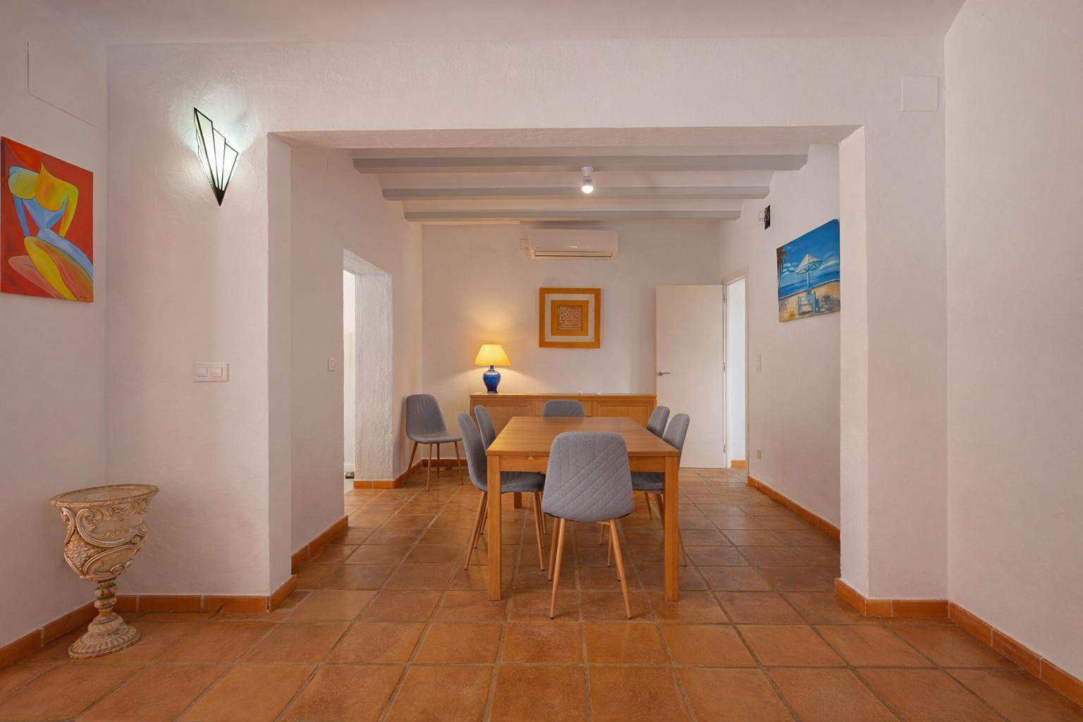 Comedor con mesa de madera y sillas tapizadas en gris, suelos de terracota y techo con vigas vistas. Incluye aire acondicionado y acceso a estancias contiguas.
