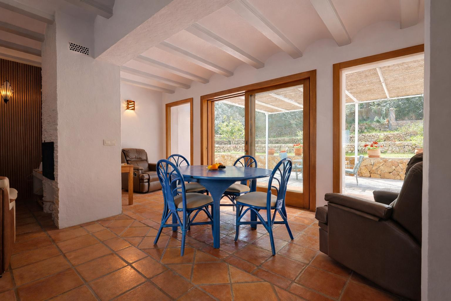 Comedor luminoso con mesa azul, vigas vistas blancas y suelo de terracota. Grandes ventanales de madera conectan el interior con el jardín y el porche exterior.