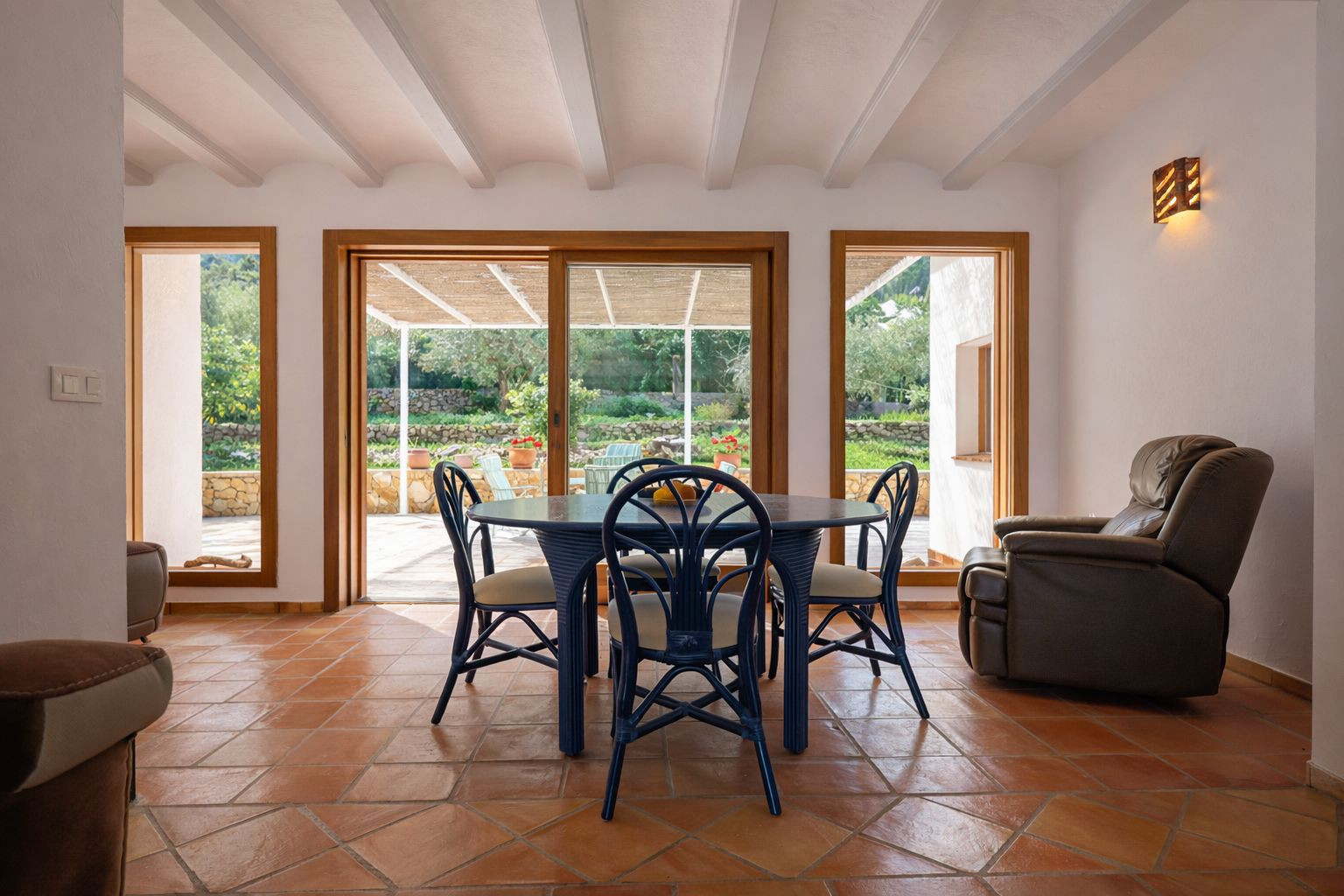 Comedor luminoso con vigas vistas blancas, suelo de terracota y acceso directo a la terraza cubierta a través de grandes ventanales de madera.