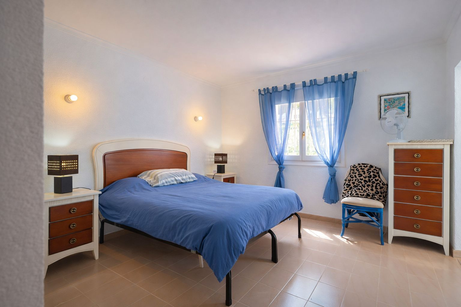 Dormitorio luminoso con suelos de gres, cama doble con cabecero de madera y mobiliario a juego. Dispone de cómoda alta y luz natural a través de cortinas azules.