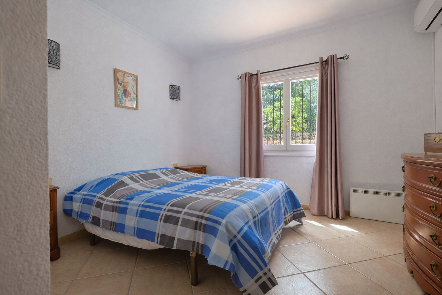 Dormitorio luminoso con suelos de gres, mobiliario de madera clásica y ventana al exterior. Incluye aire acondicionado, calefacción eléctrica y vistas al jardín.
