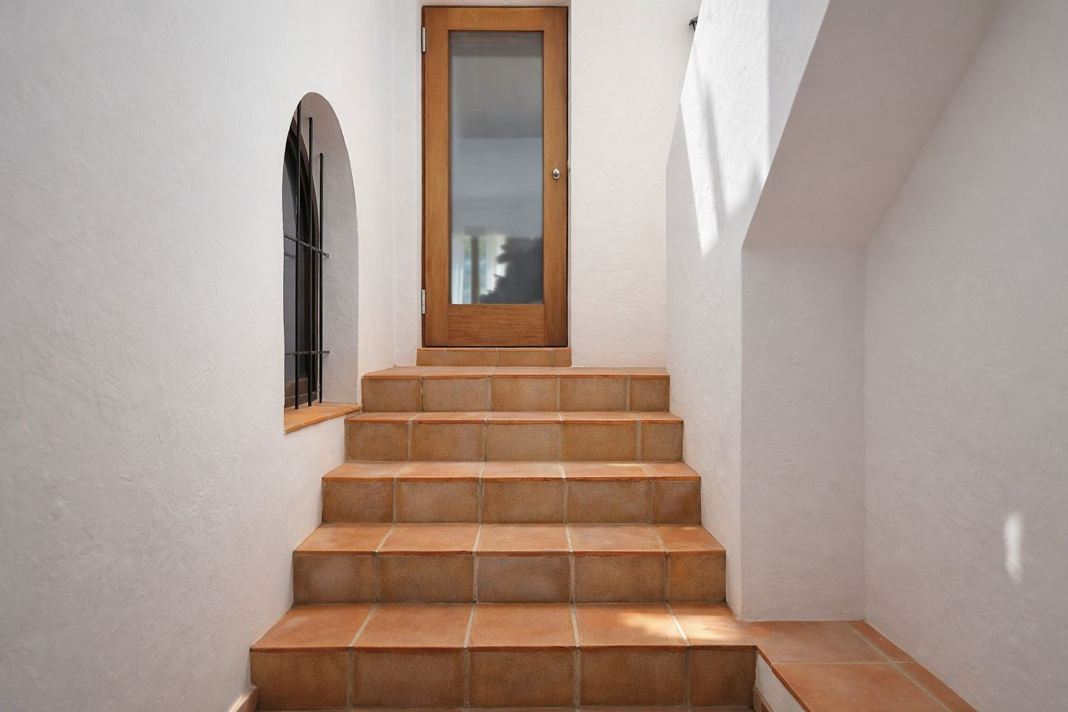 Escalera con peldaños de barro cocido que conduce a una puerta de madera y cristal, flanqueada por un arco con reja de forja y paredes blancas.