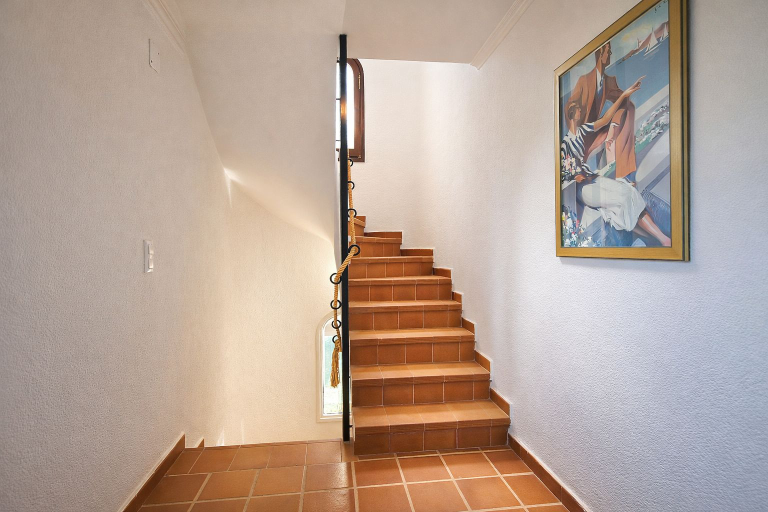 Escalera de baldosas de terracota con barandilla de hierro forjado y detalle de cuerda, iluminada por luz natural y decorada con arte de pared.