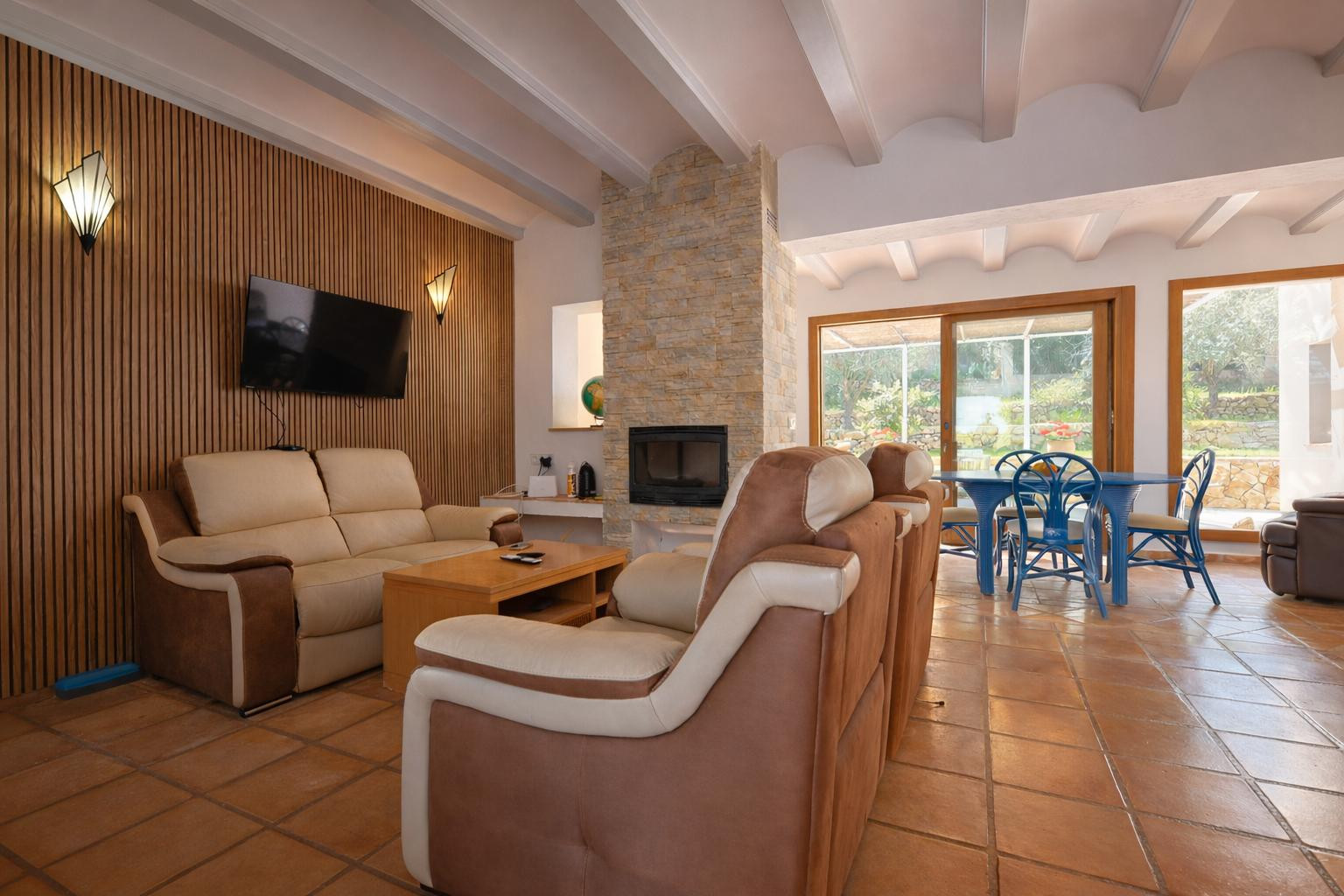 Salón-comedor con chimenea de piedra, techos de vigas blancas y suelos de terracota. Destaca la pared panelada en madera y el acceso directo al porche mediante ventanales.