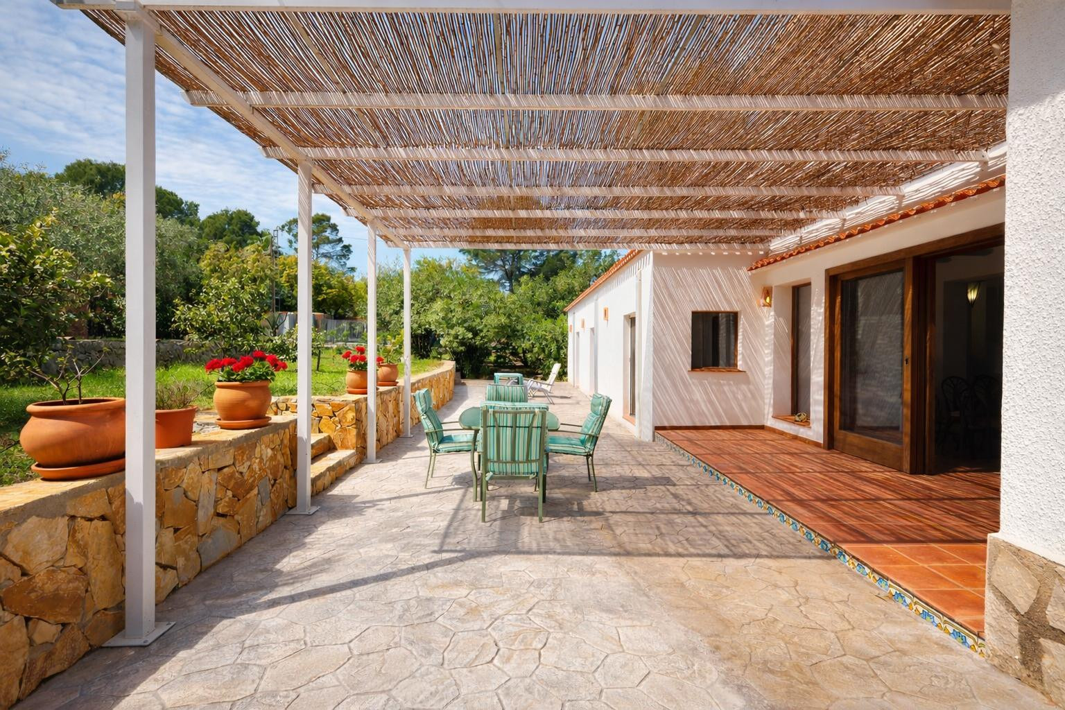 Terraza exterior con suelo de piedra y pérgola de cañizo sobre estructura metálica. Incluye zona de comedor, muros de piedra natural con macetas y acceso a la vivienda.