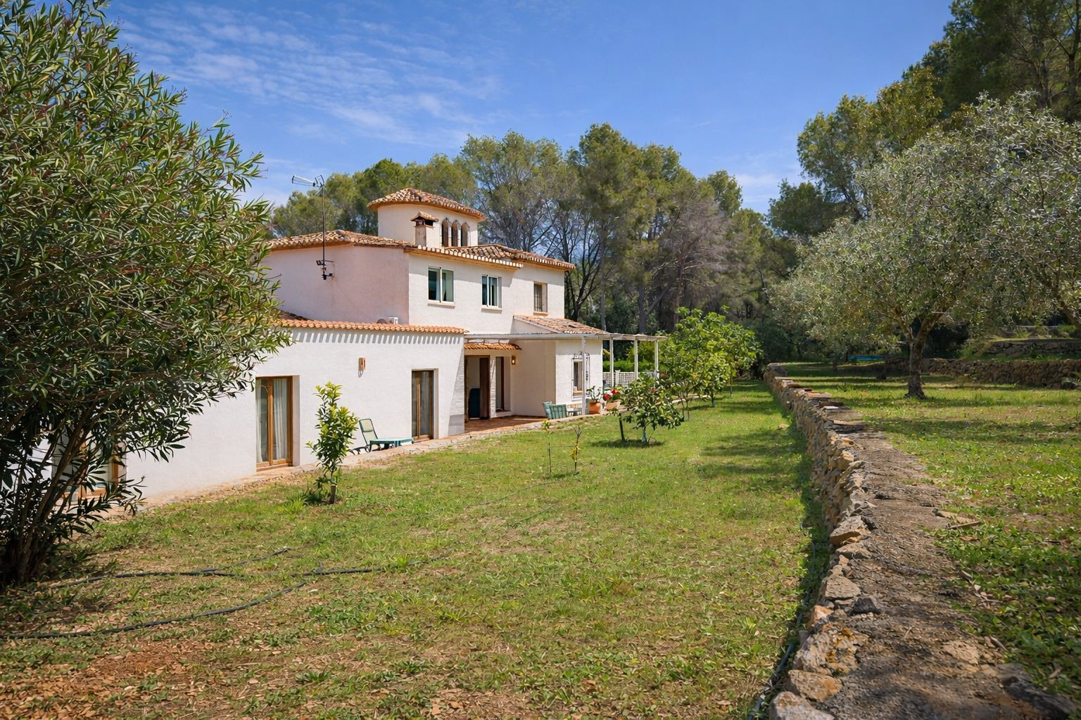 Vista exterior de la villa de dos plantas con fachada blanca y tejado de teja, rodeada de un jardín con árboles frutales y muros de piedra natural.