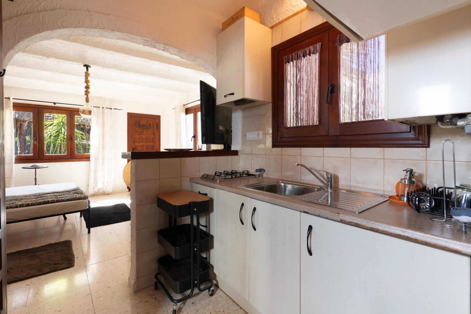 Cocina compacta de estilo rústico con armarios blancos y ventana de madera, abierta a un dormitorio luminoso mediante un arco tradicional.