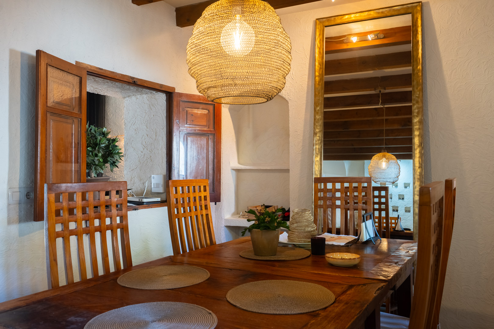 Comedor rústico con mesa de madera maciza, sillas de respaldo alto y lámpara de fibra natural. Destacan las vigas expuestas y un gran espejo con marco dorado que aporta amplitud.