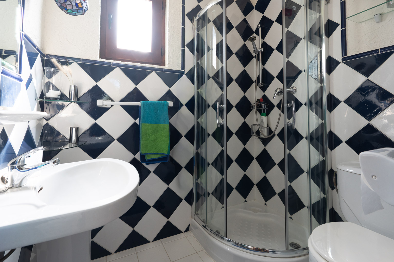 Cuarto de baño con azulejos en damero azul y blanco, equipado con una cabina de ducha de esquina curva, lavabo de pedestal blanco y ventana de madera.