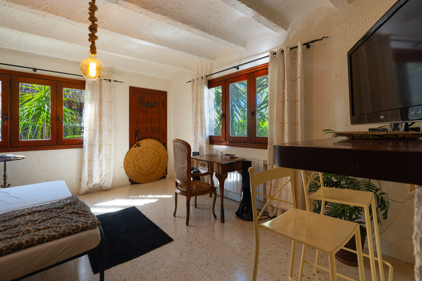 Habitación rústica con techos de vigas blancas, escritorio de madera clásica y ventanales con marcos oscuros que ofrecen vistas al jardín mediterráneo.