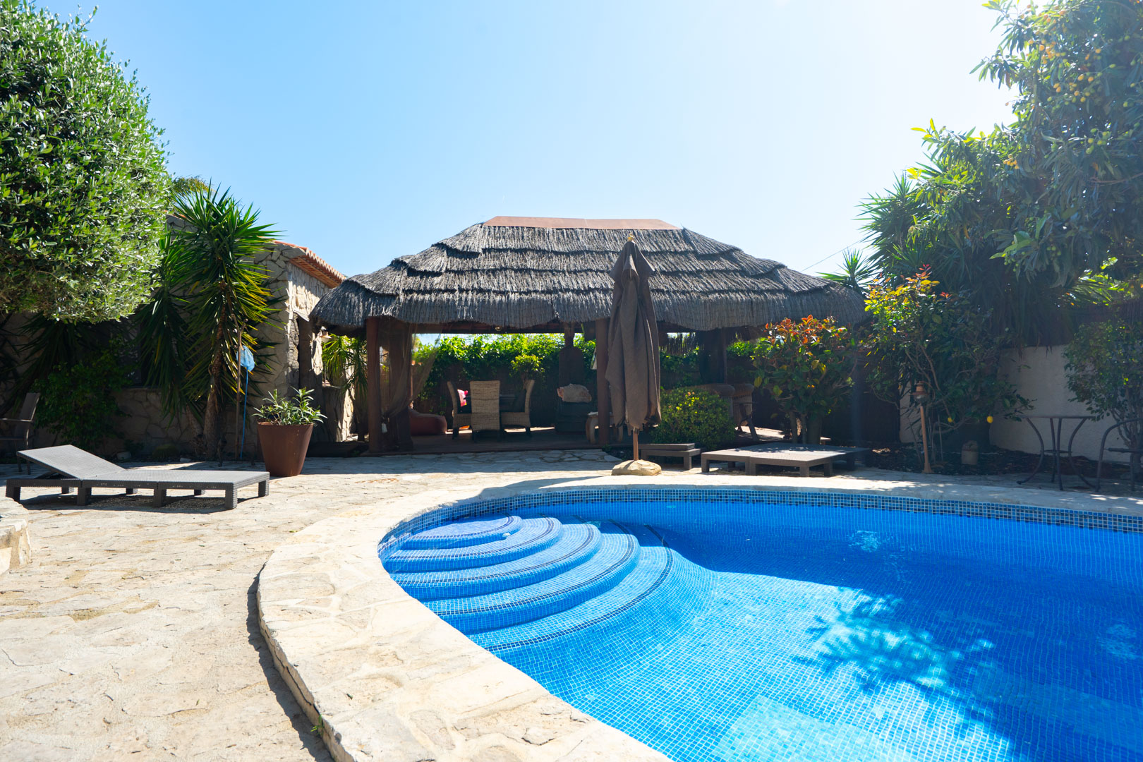 Piscina exterior rodeada de una terraza de piedra natural, con una pérgola balinesa de techo de brezo y zona de descanso entre vegetación mediterránea.