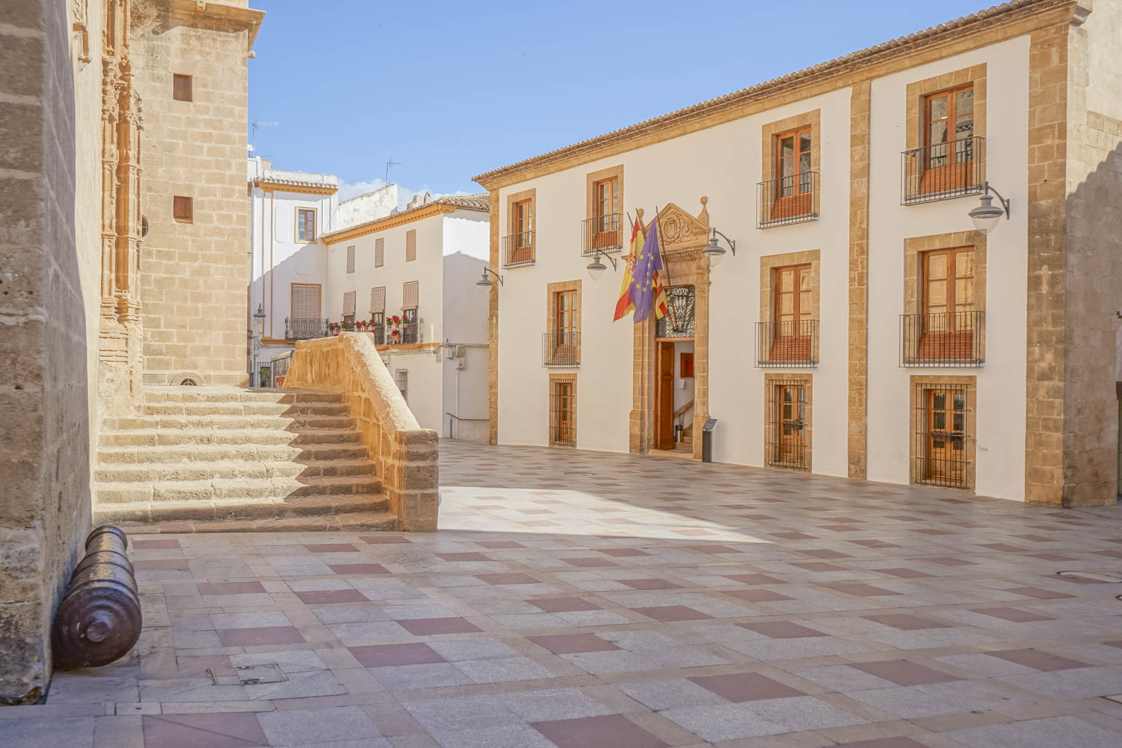 Plaza histórica en el centro de Jávea con arquitectura de piedra tosca, el ayuntamiento con banderas y la iglesia fortificada de San Bartolomé.