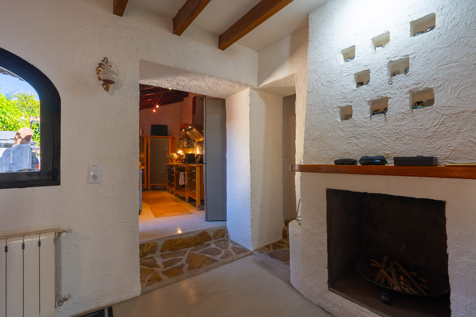 Salón rústico con vigas de madera, chimenea de obra con nichos decorativos y acceso a la cocina a través de un umbral con suelo de piedra natural.