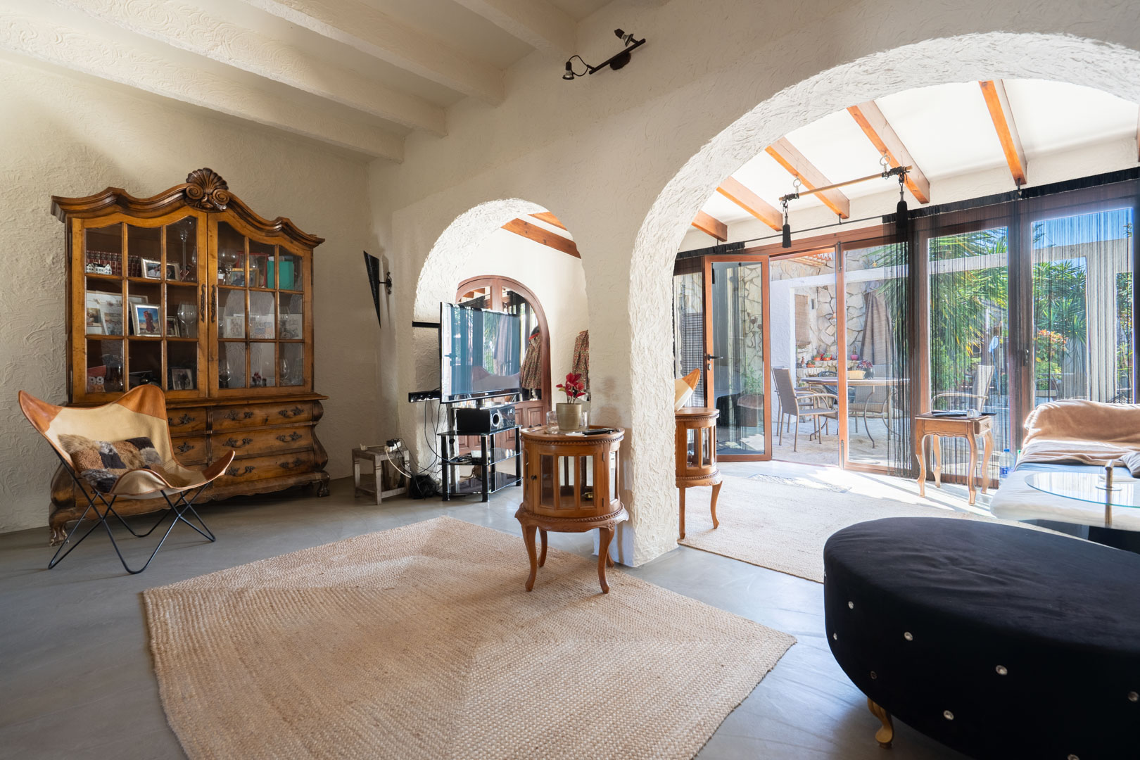 Salón rústico con vigas vistas blancas y arcos que conectan con la naya. Destacan la vitrina de madera clásica, el suelo cerámico y la abundante luz natural del patio.