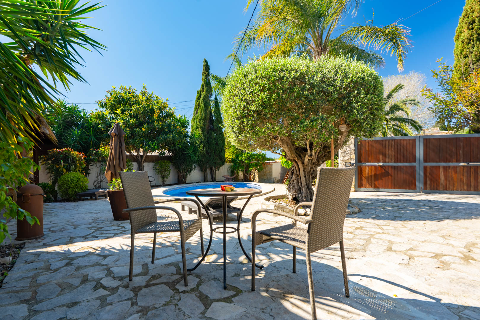 Terraza exterior con pavimento de piedra natural, mobiliario de ratán y vistas hacia la piscina privada rodeada de vegetación mediterránea y un olivo ornamental.